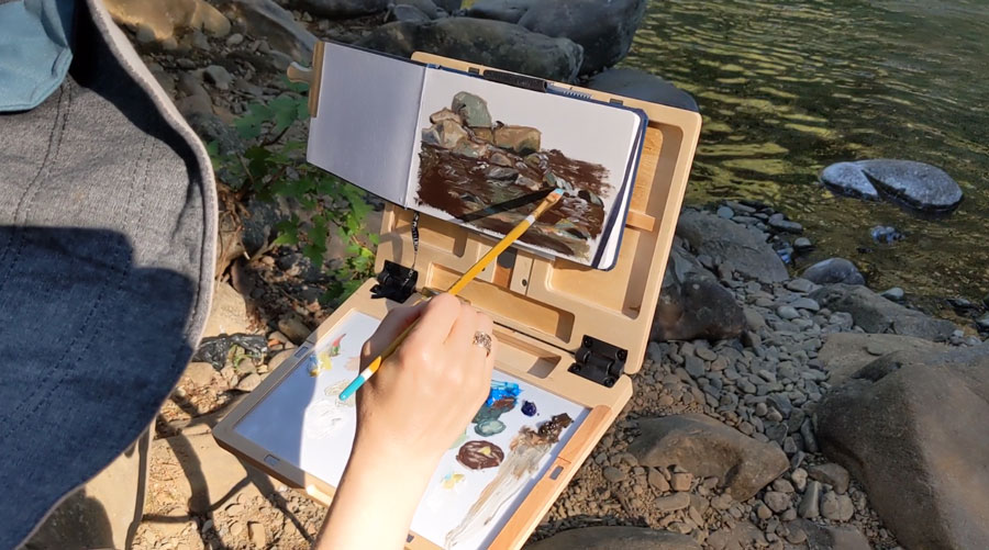 artist sitting by a stream facing an easel and painting in a hardbound sketchbook